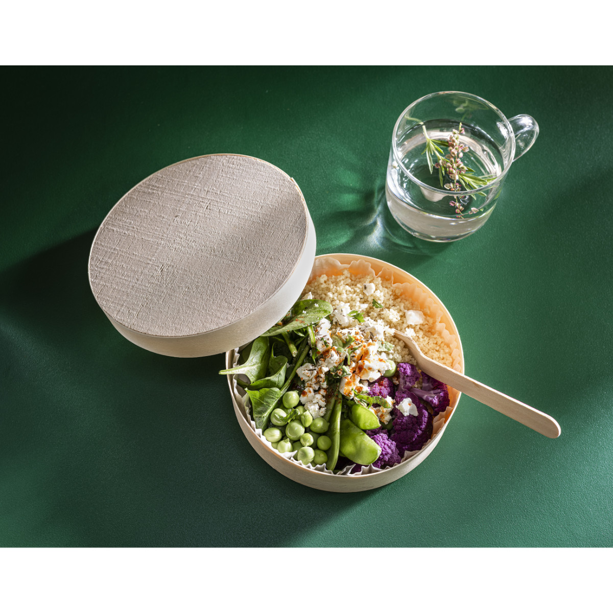 Drevená krabica na šalát | Furodet Emballages Otvorená okrúhla béžová obedárová krabička s farebným šalátom (zelenina, quinoa, fialový karfiol, hrášok) s drevenou lyžicou na zelenom povrchu, vedľa pohára vody.