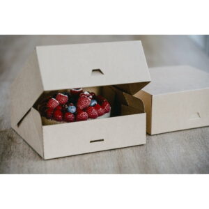 Open cardboard box reveals a berry-topped cake with raspberries and blueberries on a wooden surface.