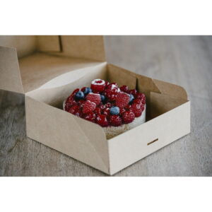 Small round cake topped with raspberries and blueberries in an open cardboard box on a wooden table.