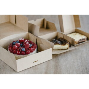 Open cardboard bakery boxes displaying assorted desserts on a wooden table, foreground berry-topped cake with raspberries and blueberries.